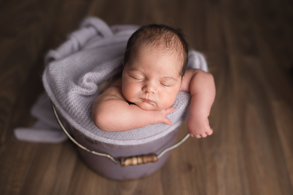Sleepy Newborn Baby Girl posed in a purple basket with her arm drapped over the side