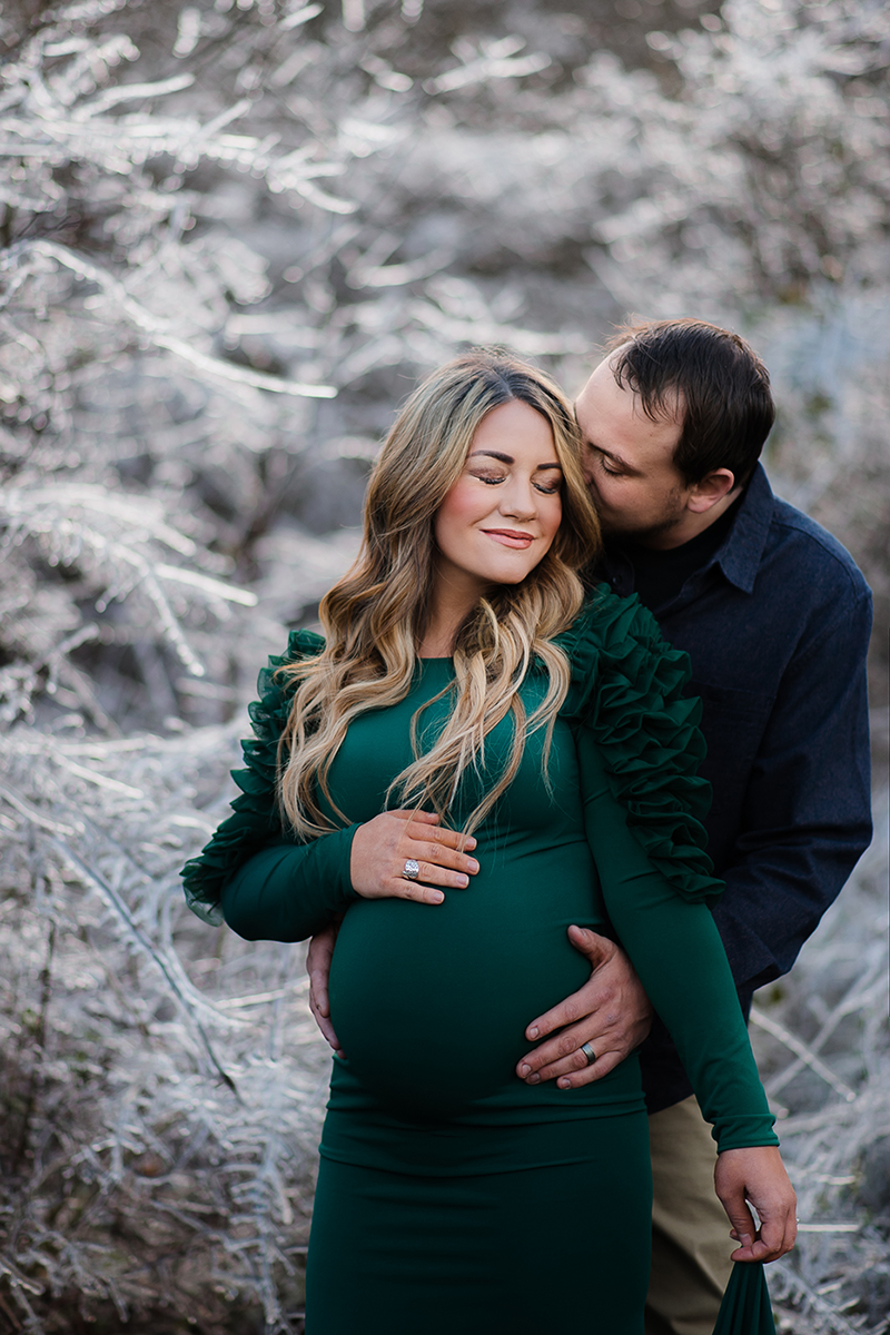 Pregnant woman in green dress, standing Infront of icy trees with husband kissing her head