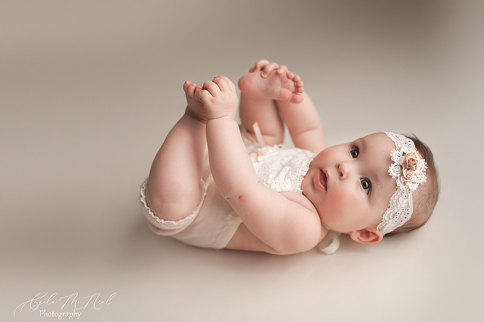 Small newborn baby, wearing a cream colored romper and lace headband, holding her toes while laying on her back