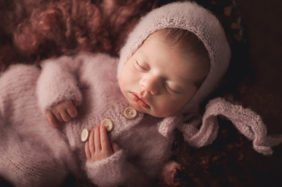 Newborn Baby Girl sleeping on dark pink wool blanket wearing a pink knit romper and a pale pink bonnet