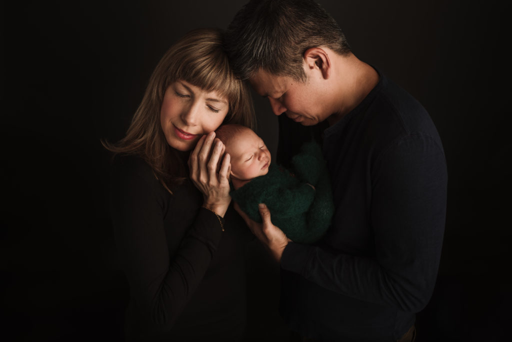 mom with eyes closed, holding newborn baby while dad is looking at sleeping baby