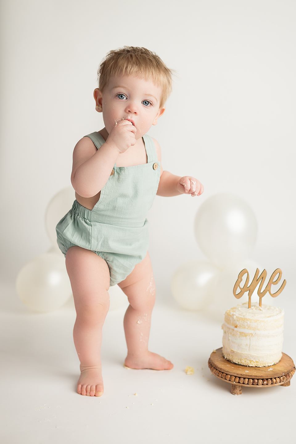 Little boy in sage romper standing above a cake that says one