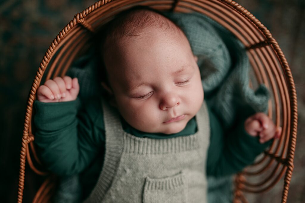 Sleeping baby laying in a boho wicker basket with sage green knit overalls