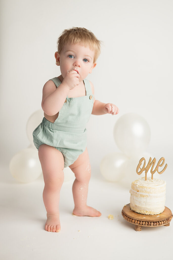 First Birthday Celebration with Little boy standing up, wearing a periwinkle bubble romper. Simple White Naked cake with golden word ONE in cursive