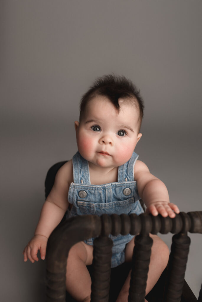 Dark haired Baby wearing jean overalls looking towards the camera sitting on a black spindle bed