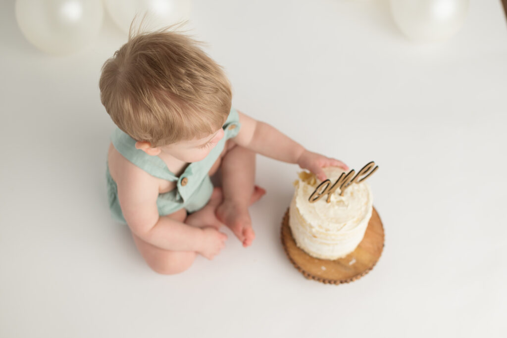 blond haired baby wearing a seafoam green romper touching a small birthday cake with a gold one on it.