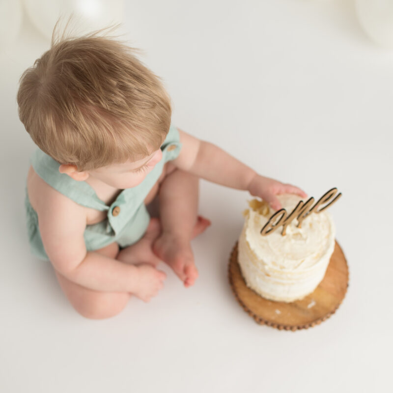 blond haired baby wearing a seafoam green romper touching a small birthday cake with a gold one on it.