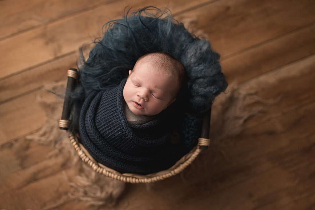 Sleeping newborn baby in a navy blue wrap, laying in a brown basket with boho backdrop