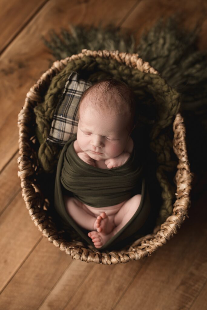 sleeping newborn baby posed in a boho basket wrapped in a green wrap and plaid pillow