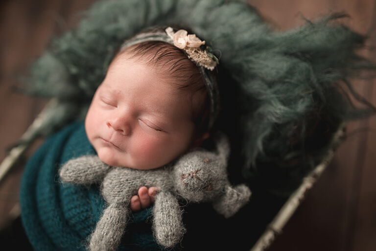 newborn baby swaddled in a jewel green wrap, sleeping with her hands out holding a stuffed bunny