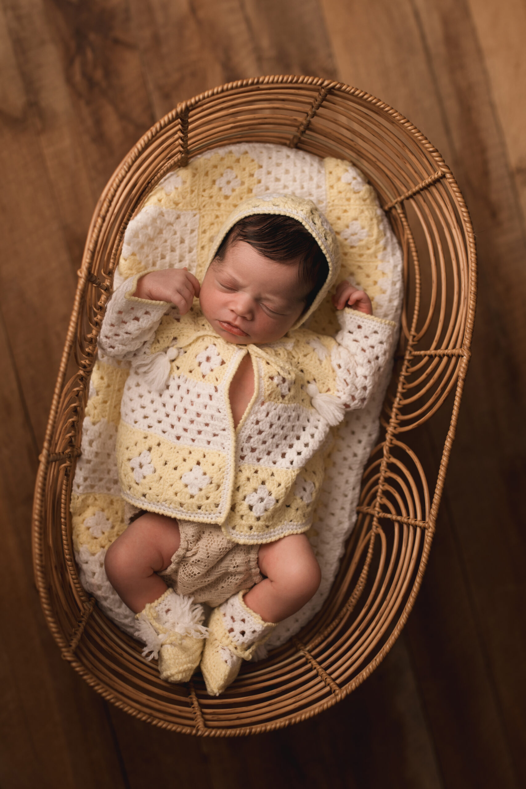 Newborn Baby Girl laying in a rattan crib, wearing a vintage yellow crochet sweater with bonnet and slippers